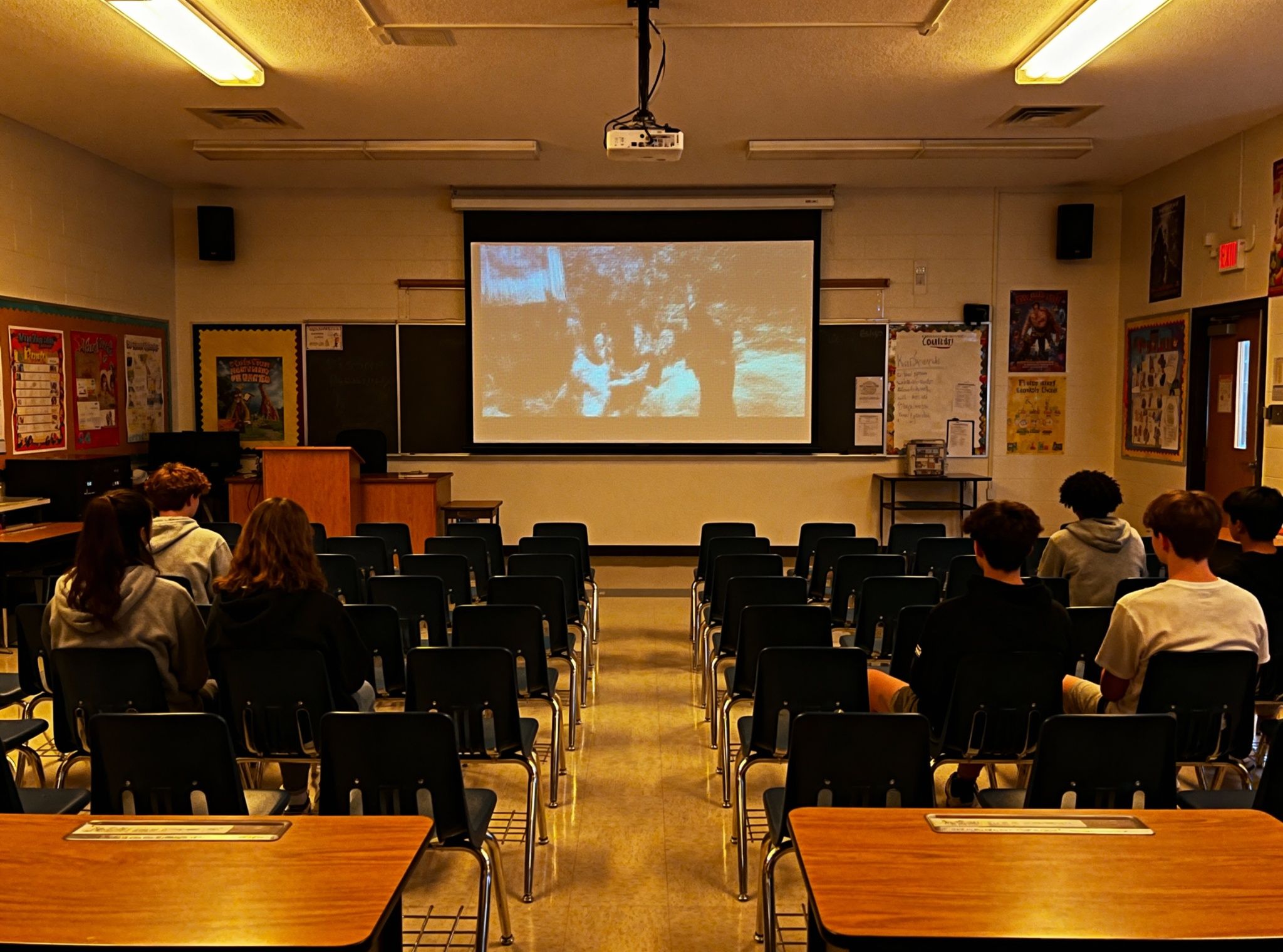 Students sitting in a classroom watching a movie projected on a large screen at the front of the room.